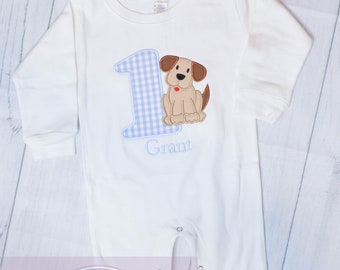 May include: White long-sleeve onesie with a blue and white gingham number one, a brown dog with a red nose, and the name "Grant" embroidered on the front.
