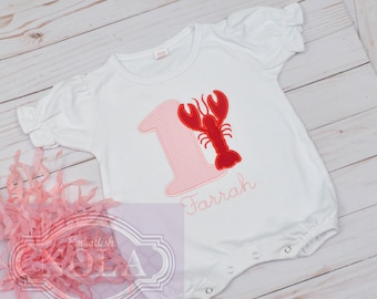 May include: White onesie with a pink and red embroidered number one and a red lobster. The onesie has the name "Farrah" embroidered below the number.