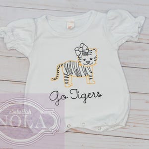 May include: White baby romper with short, ruffled sleeves. The romper features an embroidered tiger with a bow and the words "Go Tigers" in black script. The tiger has black stripes and yellow accents.