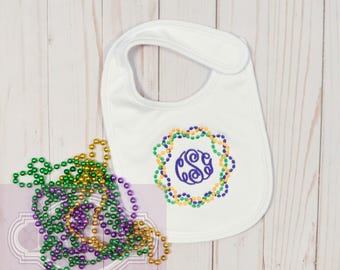 May include: White baby bib with a purple monogram surrounded by a wreath of green, gold, and purple beads. The bib is on a white wooden surface.