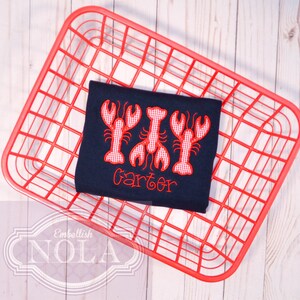 May include: A navy blue t-shirt with three red and white gingham embroidered lobsters and the name "Carter" embroidered below.
