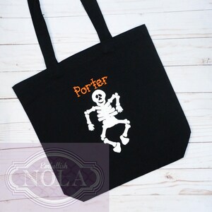 May include: Black tote bag with a white skeleton design and the name "Porter" in orange. The bag has two black handles and is made of a sturdy fabric. The skeleton is dancing, making it a fun Halloween accessory.