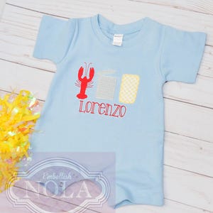 May include: A light blue baby onesie with a red lobster, a checkered box, and a yellow and white checkered box. The onesie is embroidered with the name "Lorenzo".
