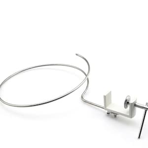 May include: A silver-colored metal clamp with a white base and a curved wire. The clamp has a screw mechanism and a handle. The wire forms a circular shape. The object is on a white background.