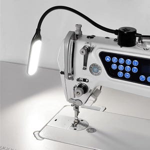 Puede incluir: Máquina de coser industrial blanca con una luz LED negra flexible. La luz ilumina la aguja y el área de trabajo. La máquina tiene un panel de control digital con botones azules.