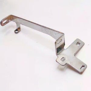 May include: A silver-colored metal mounting bracket with a bent, angular design. The component features multiple flat sections with pre-cut elongated slots for fasteners, showing a metallic finish with visible surface texture and industrial edges.