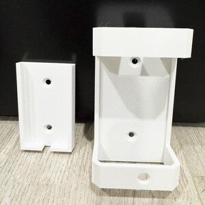 May include: Two white 3D-printed wall mounts. One is rectangular with a slot at the bottom, the other is a larger, open-front design. Both have circular holes for mounting hardware. The mounts are designed to hold an item.