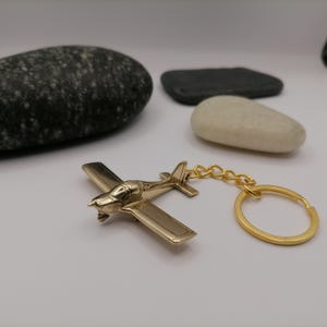 Op de afbeelding: Een goudkleurige vliegtuig sleutelhanger met een gouden ketting en ring. Het miniatuurvliegtuig heeft gedetailleerde vleugels, een cockpit en een propeller. De sleutelhanger is een decoratief accessoire.