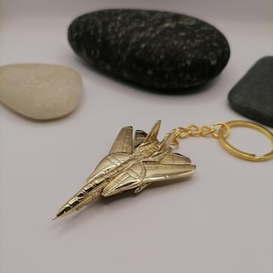 Könnte beinhalten: Ein goldfarbener Metall-Flugzeug-Schlüsselanhänger. Der Schlüsselanhänger zeigt ein detailliertes Miniatur-Flugzeugdesign mit Flügeln und einem Heck. Der Schlüsselanhänger ist an einer goldfarbenen Kette und einem Ring befestigt. Der Hintergrund umfasst glatte Steine.