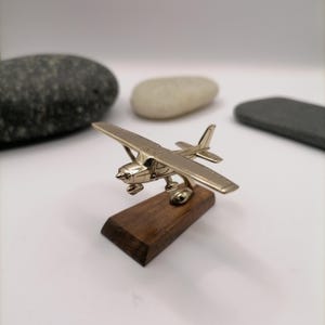 Pode incluir: Um pequeno modelo de avião em metal prateado polido com design de asa alta, hélice e trem de pouso. A aeronave em miniatura está montada sobre uma base retangular de madeira castanho-escura, apresentando um acabamento metálico detalhado.