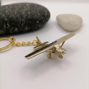 Könnte beinhalten: Ein goldfarbener Flugzeug-Schlüsselanhänger mit detailliertem Design. Der Schlüsselanhänger zeigt ein kleines Propellerflugzeug mit Flügeln und einem Heck. Das Flugzeug ist an einer goldenen Kette und einem Schlüsselring befestigt. Der Hintergrund umfasst einen schwarzen Stein und einen weißen Stein.