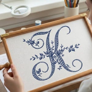 May include: A wooden embroidery hoop holds a white fabric with a detailed blue embroidered letter "K". The letter is adorned with floral and leaf patterns. The hoop has brass hardware and a wooden handle.