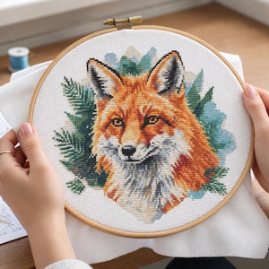 May include: A circular wooden embroidery hoop containing a cross-stitch project on white fabric. The design features a detailed portrait of a fox with orange, white, and black fur, surrounded by stylized green pine needles and blue foliage accents.