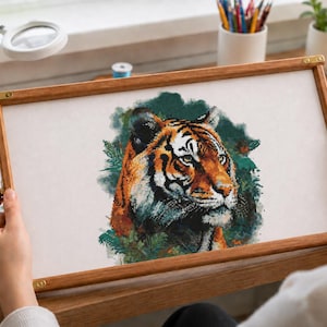 May include: A rectangular wooden embroidery frame holding a white fabric panel featuring a detailed, colorful cross-stitch or needlepoint portrait of a tiger. The tiger is depicted in shades of orange, black, and white, surrounded by green foliage.