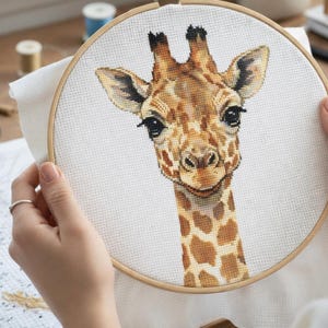 Op de afbeelding: Kruissteekborduurwerk van een giraffekop en -hals, met bruine en bruinbeige vlekken op een witte achtergrond. De giraffe heeft donkere ogen en een glimlachende uitdrukking. De borduursel zit in een houten hoepel.
