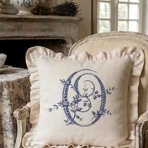 Peut inclure: Un coussin crème avec la lettre « O » brodée en bleu et des motifs floraux. Le coussin est bordé d'une volute et posé sur une chaise vintage. L'ensemble dégage une atmosphère classique et élégante, idéale pour la décoration intérieure.