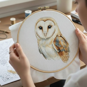 May include: A cross-stitch embroidery hoop featuring a detailed watercolor image of a barn owl. The owl has a heart-shaped face, dark eyes, and brown and white feathers. The hoop is held by a person, with craft supplies visible in the background.