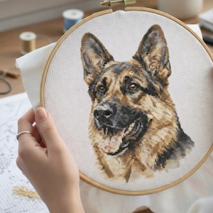 May include: A cross-stitch embroidery of a German Shepherd dog in a wooden hoop. The dog's face is detailed with brown, black, and tan thread, capturing its alert expression. The craft project is in progress, with supplies visible in the background.