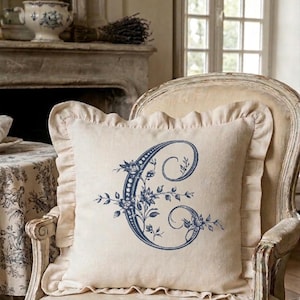 Puede incluir: Un cojín cuadrado color crema con borde con volantes, con una gran letra "C" azul adornada con elementos florales. El cojín está colocado sobre una silla de estilo vintage. Ambiente clásico y elegante.