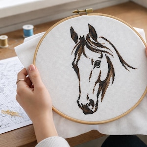 May include: A circular wooden embroidery hoop holding a piece of white fabric with a cross-stitch design of a horse head. The horse is rendered in shades of dark brown and black thread, showing the profile of the animal with a flowing mane.