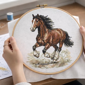 Puede incluir: Bordado de punto de cruz de un caballo marrón con cara y patas blancas, en un aro de madera. El caballo está representado a medio galope, con una melena y cola negras y fluidas. El fondo es de color beige claro.