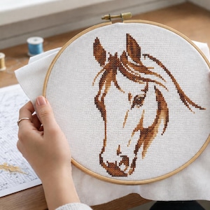 Puede incluir: Un bastidor de bordado de punto de cruz con un diseño de cabeza de caballo marrón sobre tela blanca. El bastidor de madera está sostenido por manos, con un patrón y materiales de costura visibles. Un proyecto artesanal.