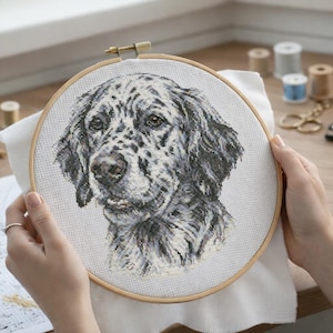 Pode incluir: Um bastidor de bordado com um retrato detalhado de um cão em ponto cruz. O cão tem uma pelagem manchada em preto e branco. O fundo inclui carretéis de linha e outros materiais de artesanato.
