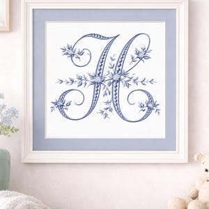 以下が含まれることがあります： 青い花の刺繍が施された文字「H」が描かれた額装されたアートワーク。 文字は花のアクセントで囲まれ、ライトブルーのマットが付いた白いフレームに額装されています。 アートワークは明るい色の木製の表面に表示されています。