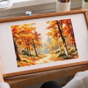 Op de afbeelding: Een rechthoekig houten borduurraam met een stuk stof waarop een landschap is afgebeeld. De afbeelding toont een pad dat door een bos slingert, gevuld met levendig oranje, geel en rood herfstgebladerte en hoge boomstammen.