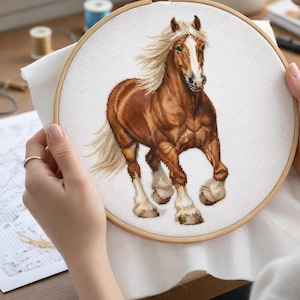 Può includere: Ricamo a punto croce di un cavallo marrone con criniera e coda bianche fluenti, posto in un telaio di legno. Il cavallo è raffigurato in posa di corsa, mostrando la sua corporatura muscolosa. Lo sfondo è in tessuto bianco.