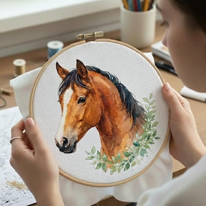 Op de afbeelding: Een borduurring met kruissteek met een gedetailleerd aquarelportret van een paardenkop. Het paard heeft een bruine vacht, een zwarte manen en een witte bles. Groene bladeren omringen de paardenkop, tegen een witte stoffen achtergrond.
