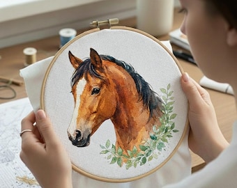 Op de afbeelding: Een borduurring met kruissteek met een gedetailleerd aquarelportret van een paardenkop. Het paard heeft een bruine vacht, een zwarte manen en een witte bles. Groene bladeren omringen de paardenkop, tegen een witte stoffen achtergrond.