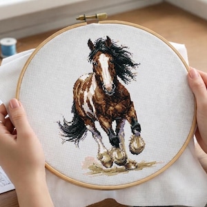 May include: A cross-stitch embroidery hoop featuring a brown and white horse in mid-gallop. The horse has a black mane and tail, and is set against a white background. The wooden hoop is held by hands.