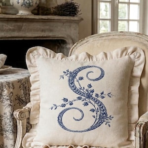 Pode incluir: Uma almofada quadrada creme com uma letra "S" bordada floral azul é exibida em uma cadeira creme. A almofada tem uma borda com babados. O fundo inclui uma lareira, uma mesa com uma toalha de mesa floral e uma janela.