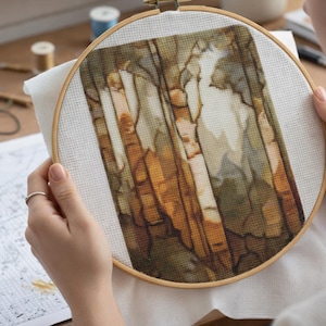 May include: A cross-stitch project in progress, showing a detailed landscape design within a wooden embroidery hoop. The artwork uses browns, greens, and whites, held by a person. The project is on a white fabric. The hoop is a common craft item.