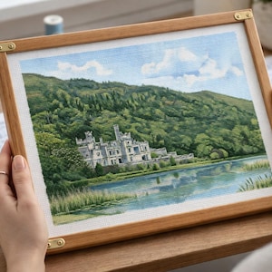 Peut inclure: Point de croix d'un paysage de château au bord d'un lac, encadré dans un cadre en bois brun clair. La scène présente un château blanc, des arbres verts et un ciel bleu nuageux. Le cadre a des coins en laiton.