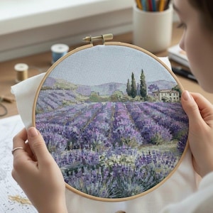 May include: A cross-stitch embroidery hoop featuring a detailed landscape of a lavender field. The scene includes rows of purple lavender, green trees, and a building under a light blue sky. The embroidery is held in a wooden hoop.