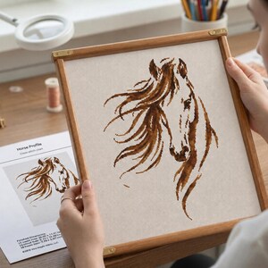 Puede incluir: Un bordado de punto de cruz enmarcado de un perfil de caballo, con una melena marrón fluida y una cara detallada. La obra de arte está enmarcada en un marco de madera. Un patrón impreso es visible debajo.