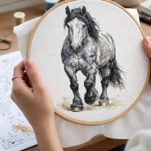 May include: A cross-stitch embroidery of a grey horse with a black mane and tail, set in a wooden hoop. The horse is depicted in a walking pose. The embroidery is held by a person.