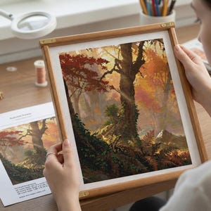 May include: A cross-stitch embroidery project in a wooden frame depicting an autumn forest scene with a large central tree, vibrant red and orange foliage, and green mossy ground. The artwork is displayed alongside a printed pattern guide.