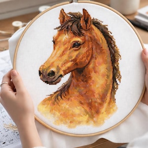 May include: A detailed cross-stitch embroidery of a horse's head, showcasing shades of brown, gold, and black. The artwork is stretched within a wooden embroidery hoop, held by hands. The craft project is on a white fabric, with a detailed design.