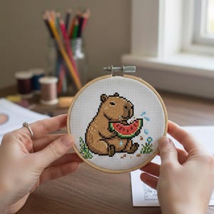 May include: A cross-stitch hoop featuring a pixelated capybara eating watermelon. The capybara is brown with a red and green watermelon slice. The hoop is held by hands, with a wooden frame and white fabric.