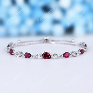 Peut inclure: Un bracelet réglable de couleur argentée orné d'une série de pierres rouge foncé taillées en poire alternant avec de petites pierres claires serties dans des maillons métalliques torsadés. Le motif central présente une pierre rouge.