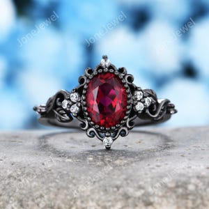 Könnte beinhalten: Ein Ring aus schwarzem Metall mit einem zentralen, oval geschliffenen, tiefroten Edelstein, der von einer dekorativen Filigranfassung umgeben ist. Die Ringschiene ist mit kleinen klaren Steinen und blattartigen Motiven verziert.