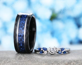 May include: A black wedding band with a blue inlay next to a silver engagement ring. The engagement ring has a central diamond and blue sapphire accents. The background is a soft blue.