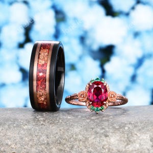 May include: Two rings: a black band with a red and gold inlay, and a rose gold ring with a large red gemstone and smaller green stones. The rose gold ring has rose details.