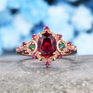 May include: A rose gold ring featuring a large oval-cut deep red center stone, flanked by two smaller round green stones set within crescent shapes, accented by multiple tiny round pink stones arranged in a decorative floral-inspired metal setting.