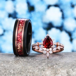 May include: A black band ring with a central inlay of red and gold flakes alongside a rose gold-toned ring set featuring a pear-shaped red gemstone surrounded by small clear stones and a matching curved band with marquise and round clear stones.