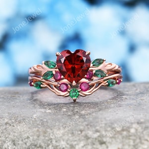 Op de afbeelding: Een roségouden ring met een centraal hartvormig rood edelsteen. De band is ontworpen met bladvormige groene stenen en ronde roze en groene accentstenen in een rankachtig patroon. Het metaal heeft een gepolijste afwerking.
