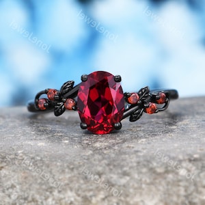 May include: A black metal ring featuring a large oval-cut deep red gemstone as the centerpiece. The band is adorned with small leaf-shaped accents and several tiny round red stones on both sides of the main setting, creating a botanical design.